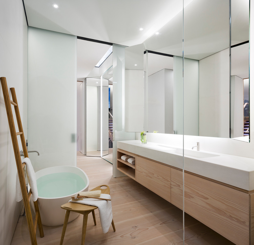 Ejemplo de cuarto de baño actual con lavabo integrado, armarios con paneles lisos, puertas de armario de madera clara, bañera exenta, paredes blancas y suelo de madera clara
