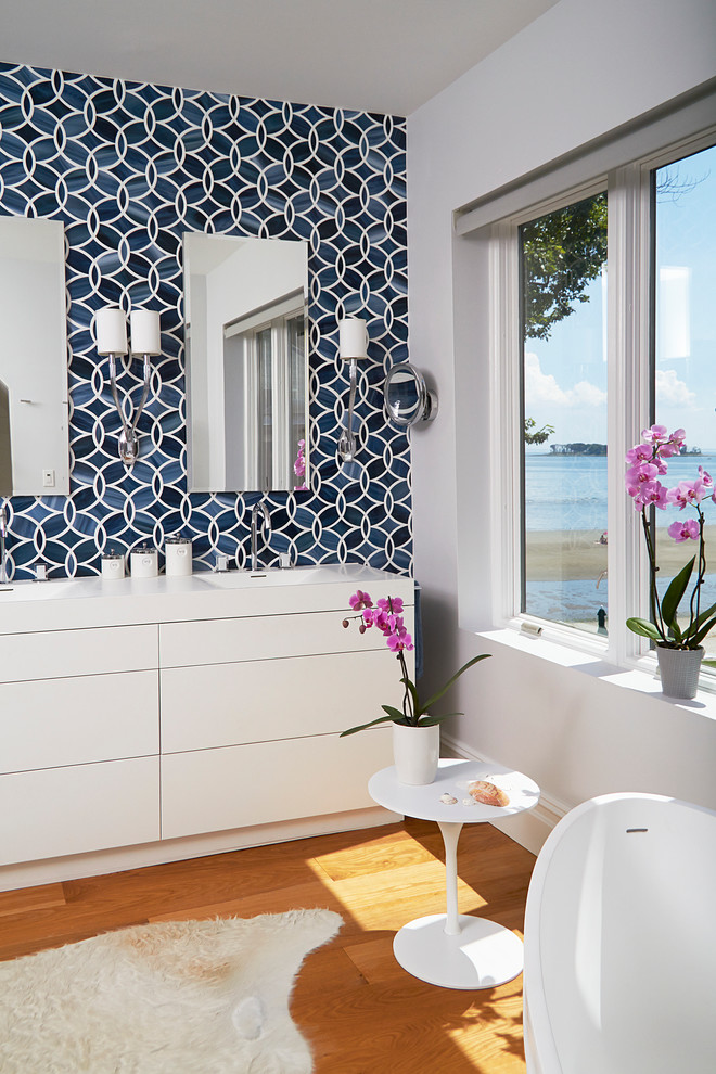Cette image montre une salle de bain design avec un placard à porte plane, des portes de placard blanches, un carrelage bleu, un mur blanc, un sol en bois brun, un lavabo intégré et du carrelage bicolore.