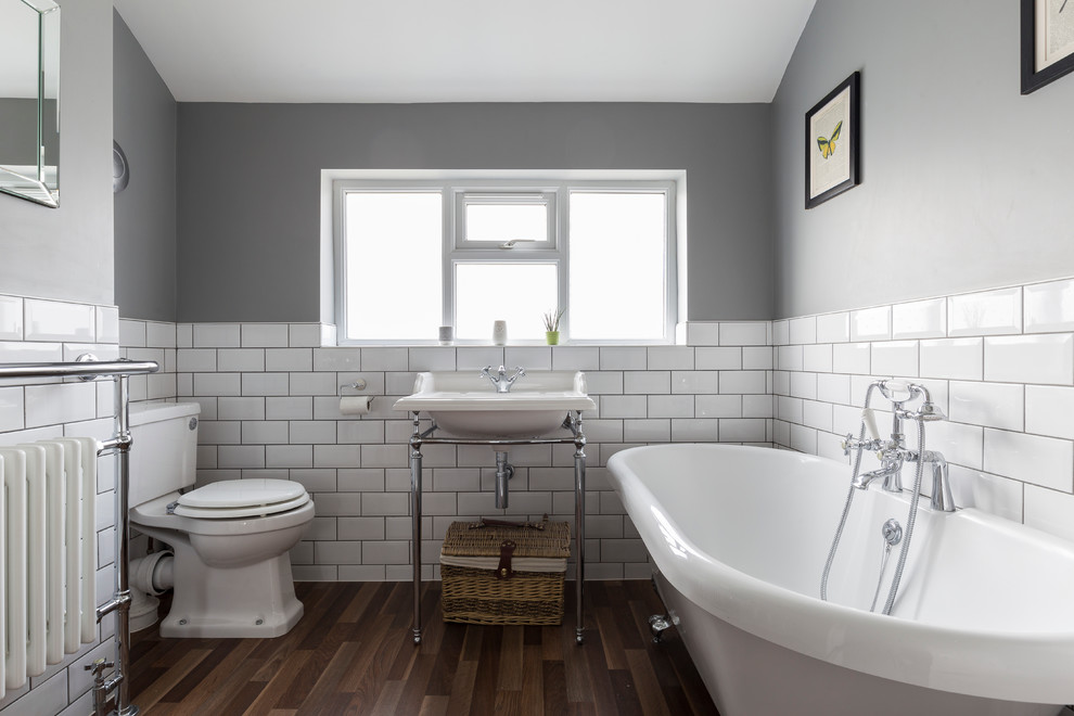 Inredning av ett industriellt badrum, med ett badkar med tassar, en toalettstol med separat cisternkåpa, vit kakel, tunnelbanekakel, grå väggar, ett konsol handfat och mörkt trägolv