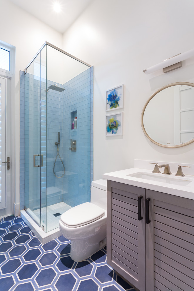Exemple d'une salle d'eau bord de mer de taille moyenne avec des portes de placard grises, une douche d'angle, un carrelage bleu, un mur blanc, un lavabo encastré, WC séparés, un sol en carrelage de porcelaine, un plan de toilette en surface solide, une cabine de douche à porte battante et un placard avec porte à panneau encastré.