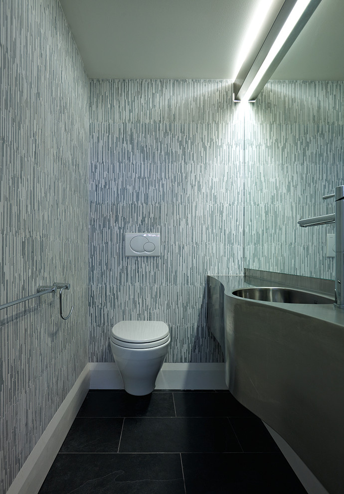 Cette photo montre une petite salle de bain principale tendance avec un lavabo intégré, un plan de toilette en acier inoxydable, WC suspendus, un carrelage noir, des dalles de pierre et un mur gris.