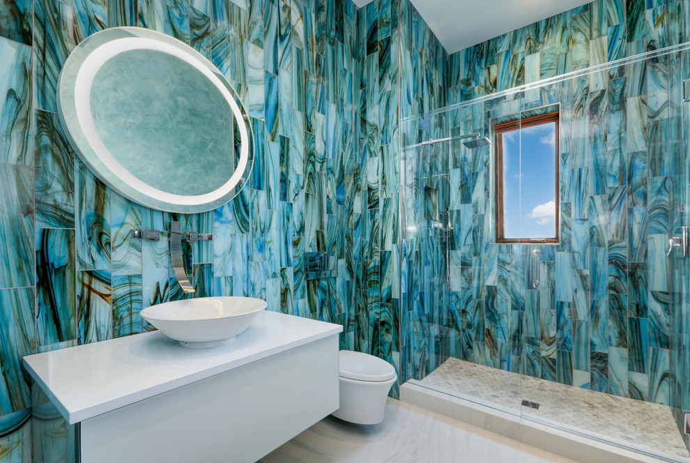 Cette photo montre une salle de bain tendance de taille moyenne avec un placard à porte plane, des portes de placard blanches, WC suspendus, un carrelage bleu, un carrelage en pâte de verre, un mur bleu, un sol en carrelage de porcelaine, une vasque et un plan de toilette en marbre.
