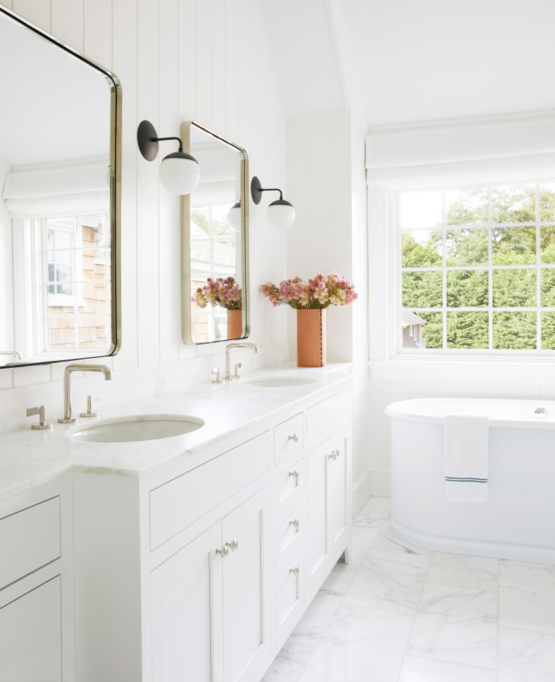 Пример оригинального дизайна: главная ванная комната среднего размера в морском стиле с белыми фасадами, белой плиткой, керамической плиткой, белыми стенами, мраморным полом, мраморной столешницей, белым полом, белой столешницей, тумбой под две раковины, встроенной тумбой, фасадами в стиле шейкер, отдельно стоящей ванной, врезной раковиной, сводчатым потолком и стенами из вагонки