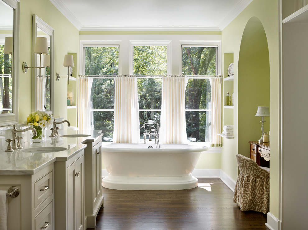 Klassisk inredning av ett en-suite badrum, med grå skåp, ett fristående badkar, gröna väggar, mörkt trägolv, ett undermonterad handfat och luckor med infälld panel