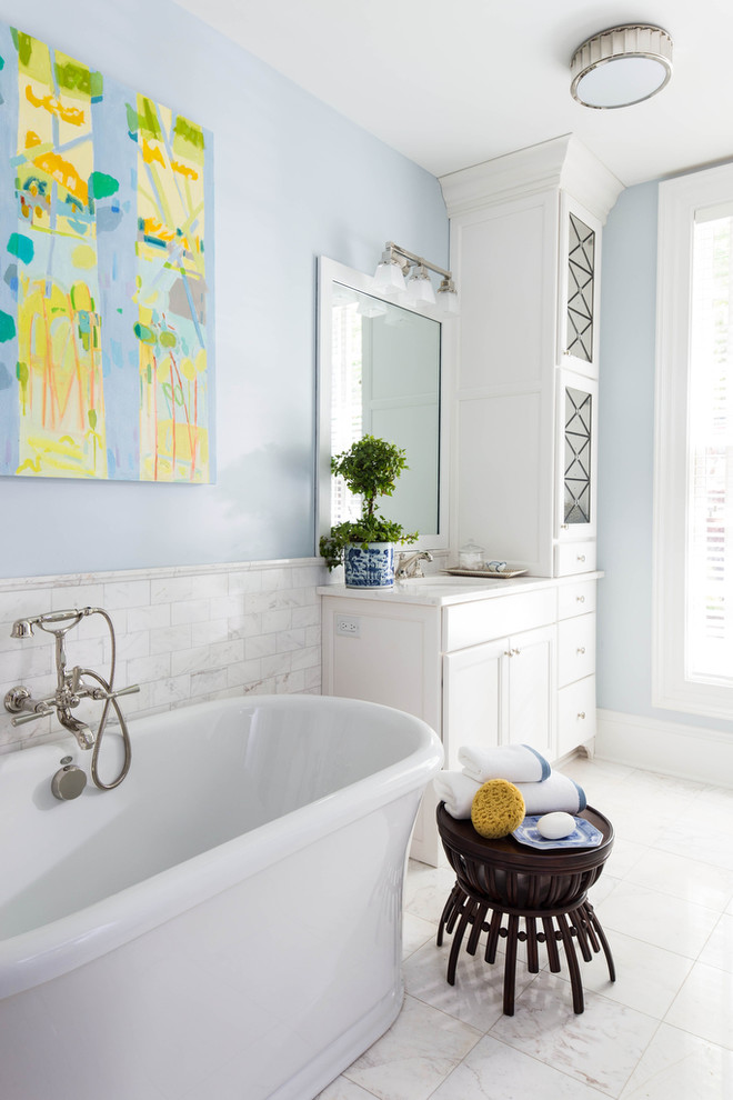Cette image montre une salle de bain rustique avec un lavabo encastré, des portes de placard blanches, une baignoire indépendante, un carrelage blanc, un carrelage de pierre, un mur bleu, un plan de toilette en marbre et un placard avec porte à panneau encastré.