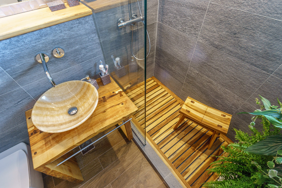 Cette image montre une petite salle de bain ethnique avec des portes de placard blanches, WC suspendus, un carrelage gris, des carreaux de céramique, un mur gris, un sol en carrelage de céramique, une vasque et un plan de toilette en bois.