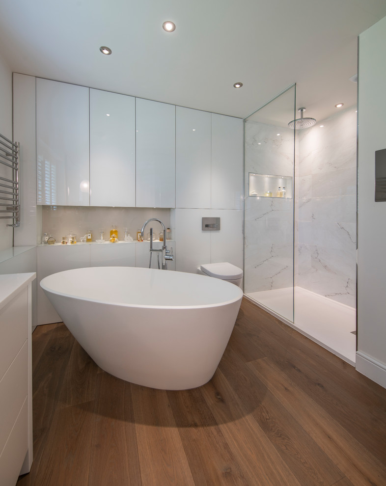 Contemporary ensuite bathroom in Surrey with white cabinets, a freestanding bath, a corner shower, a wall mounted toilet, grey tiles, white tiles, white walls, brown floors, an open shower, white worktops and flat-panel cabinets.