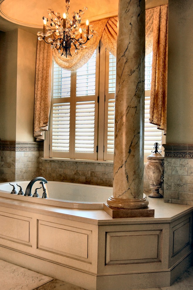 Aménagement d'une grande salle de bain principale classique avec un placard avec porte à panneau surélevé, des portes de placard beiges, une baignoire posée, un carrelage beige, des dalles de pierre, un sol en calcaire, un plan de toilette en marbre, un mur beige, un lavabo encastré et un sol marron.