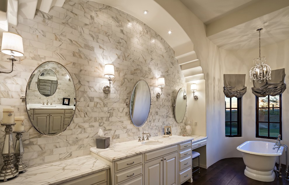 Foto på ett mellanstort medelhavsstil en-suite badrum, med luckor med upphöjd panel, vita skåp, ett fristående badkar, grå kakel, stenkakel, beige väggar, mörkt trägolv, ett undermonterad handfat och marmorbänkskiva