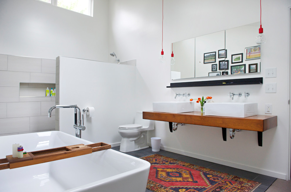 Modern inredning av ett stort en-suite badrum, med öppna hyllor, skåp i mellenmörkt trä, ett fristående badkar, en kantlös dusch, en toalettstol med hel cisternkåpa, grå kakel, porslinskakel, vita väggar, klinkergolv i porslin, ett fristående handfat, träbänkskiva, svart golv och med dusch som är öppen