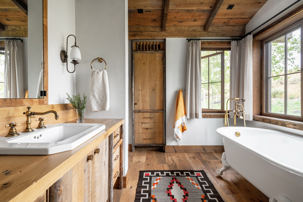Aménagement d'une salle de bain principale montagne en bois brun avec une baignoire sur pieds, un mur blanc, un sol en bois brun, un lavabo posé, un plan de toilette en bois et un placard à porte plane.