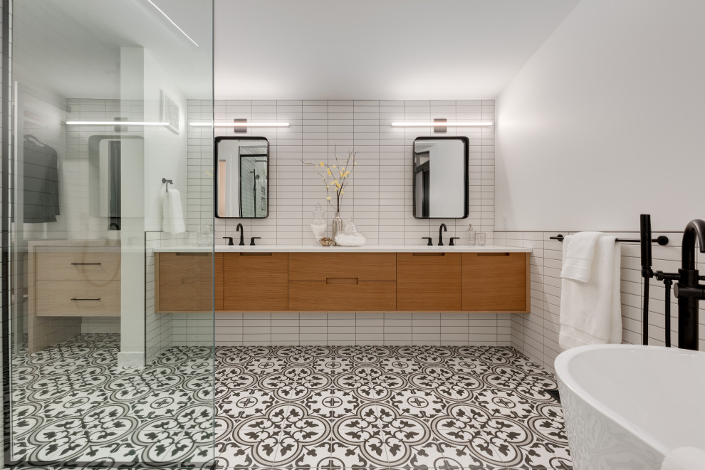 Inspiration for a contemporary ensuite bathroom in Calgary with flat-panel cabinets, medium wood cabinets, a freestanding bath, white tiles, white walls, a submerged sink, multi-coloured floors, white worktops and double sinks.
