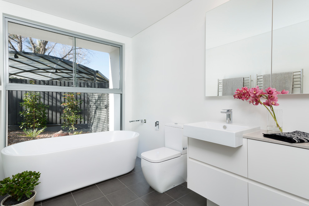 Inredning av ett modernt badrum, med släta luckor, vita skåp, ett fristående badkar, en toalettstol med separat cisternkåpa, grå kakel, vit kakel, porslinskakel, vita väggar, klinkergolv i keramik och granitbänkskiva