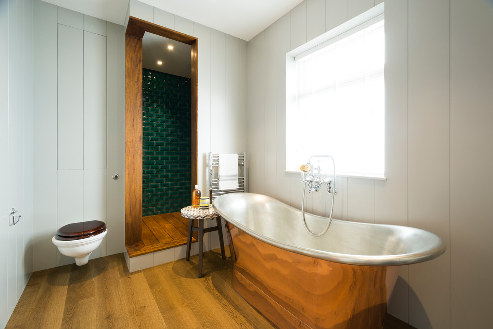 Inredning av ett lantligt badrum, med ett fristående badkar, en hörndusch, en vägghängd toalettstol, vita väggar, mellanmörkt trägolv, grön kakel och tunnelbanekakel