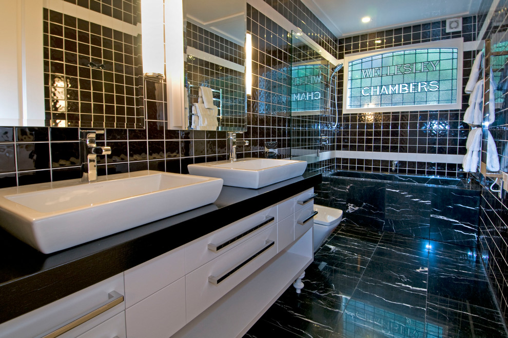 Photo of a contemporary bathroom in Auckland with flat-panel cabinets, white cabinets, an alcove bath, a shower/bath combination, a one-piece toilet, black tiles, black and white tiles, white tiles, black walls and a vessel sink.