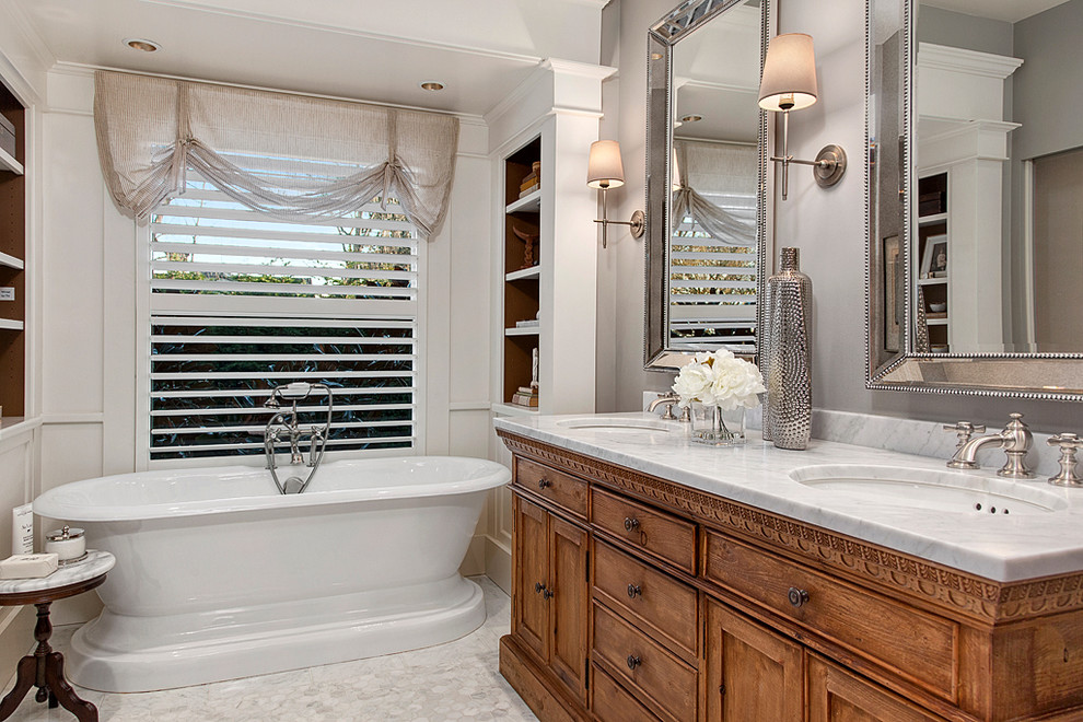 Klassisk inredning av ett mycket stort en-suite badrum, med skåp i mellenmörkt trä, ett fristående badkar, en dubbeldusch, en toalettstol med separat cisternkåpa, vit kakel, grå väggar, marmorgolv, ett undermonterad handfat, marmorbänkskiva och skåp i shakerstil