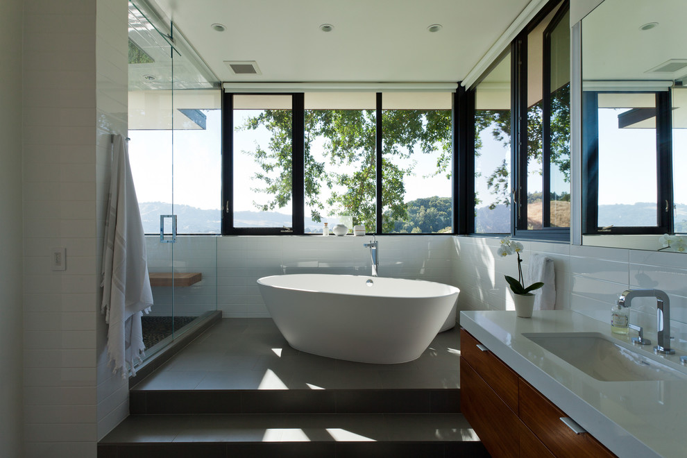 На фото: большая главная ванная комната в стиле модернизм с врезной раковиной, плоскими фасадами, фасадами цвета дерева среднего тона, отдельно стоящей ванной, душем в нише и керамогранитной плиткой