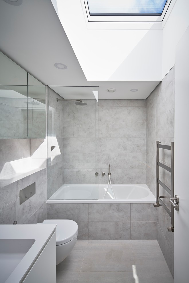 This is an example of a medium sized contemporary family bathroom in London with a built-in bath, a shower/bath combination, a one-piece toilet, grey tiles, a wall-mounted sink and tiled worktops.