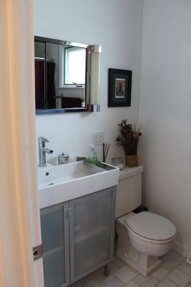 Modern inredning av ett litet badrum, med ett avlångt handfat, luckor med glaspanel, vita skåp, en toalettstol med hel cisternkåpa, stenkakel, vita väggar och marmorgolv