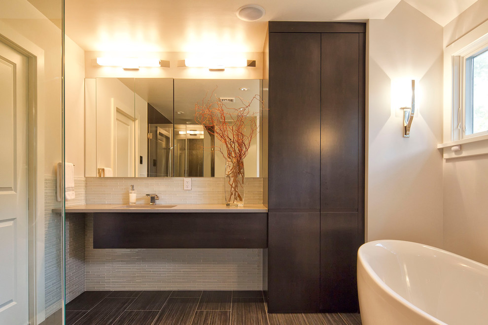 На фото: ванная комната в современном стиле с плоскими фасадами, темными деревянными фасадами, отдельно стоящей ванной, угловым душем, унитазом-моноблоком, серой плиткой, стеклянной плиткой, белыми стенами, полом из керамогранита, подвесной раковиной, столешницей из искусственного кварца, душем с распашными дверями и серой столешницей с