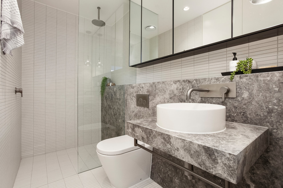 Diseño de cuarto de baño contemporáneo con armarios con paneles lisos, ducha empotrada, sanitario de pared, paredes blancas, lavabo sobreencimera, encimera de mármol, suelo blanco, ducha abierta y encimeras grises