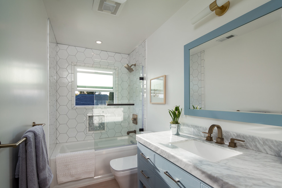 This is an example of a medium sized bohemian family bathroom in San Francisco with flat-panel cabinets, blue cabinets, an alcove bath, white tiles, white walls, light hardwood flooring, a submerged sink and marble worktops.