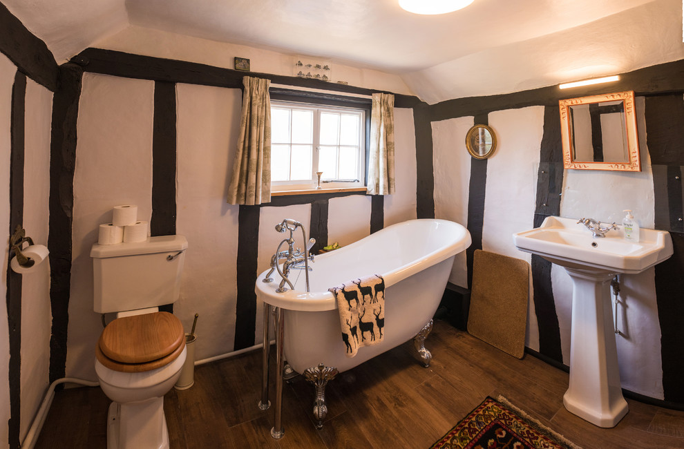 Lantlig inredning av ett litet badrum, med en toalettstol med separat cisternkåpa, ett piedestal handfat, ett badkar med tassar, flerfärgade väggar och brunt golv