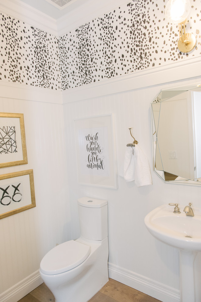 Lantlig inredning av ett badrum, med en toalettstol med hel cisternkåpa, vita väggar, ljust trägolv och ett piedestal handfat