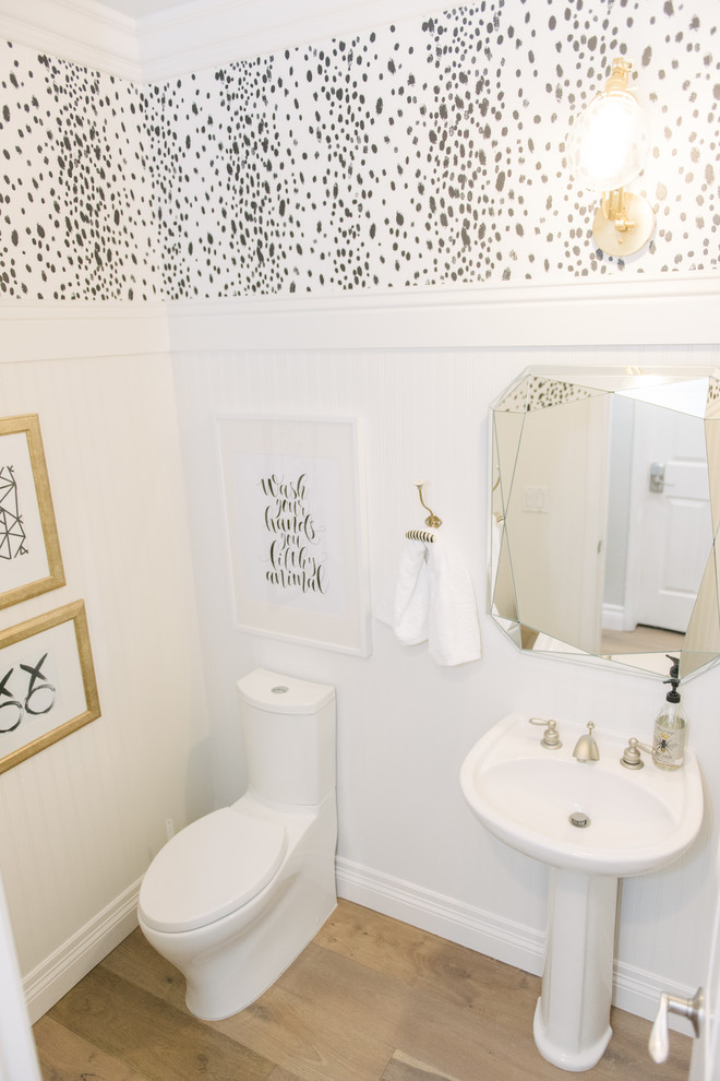 Inredning av ett lantligt badrum, med en toalettstol med hel cisternkåpa, vita väggar, ljust trägolv och ett piedestal handfat