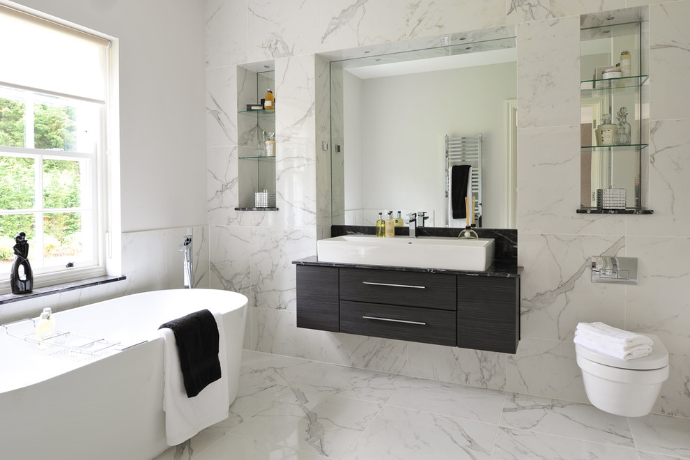 Bild på ett mellanstort funkis badrum, med släta luckor, svarta skåp, ett fristående badkar, en vägghängd toalettstol, vit kakel, ett väggmonterat handfat, porslinskakel, klinkergolv i porslin och vitt golv
