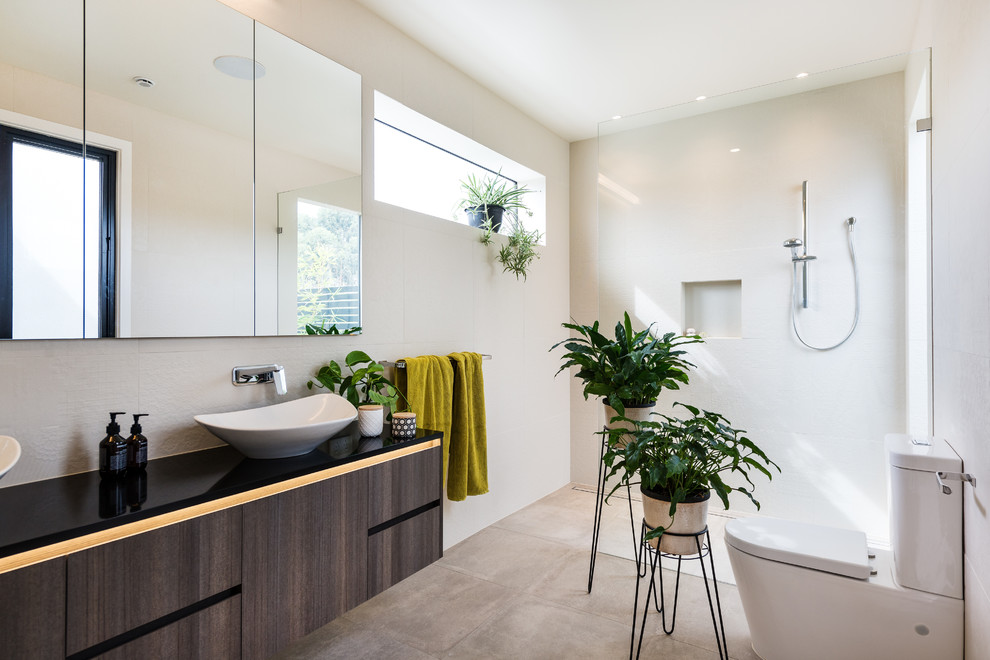 Inspiration for a modern ensuite wet room bathroom in Melbourne with dark wood cabinets, a one-piece toilet, porcelain tiles, porcelain flooring, flat-panel cabinets, white tiles, white walls, a vessel sink, engineered stone worktops, brown floors, black worktops and an open shower.