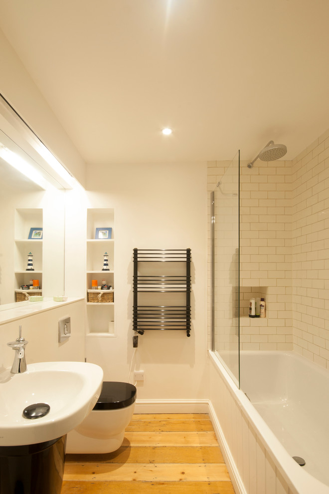 Ejemplo de cuarto de baño contemporáneo con lavabo con pedestal, bañera empotrada, sanitario de pared, baldosas y/o azulejos blancos, paredes blancas y suelo de madera clara
