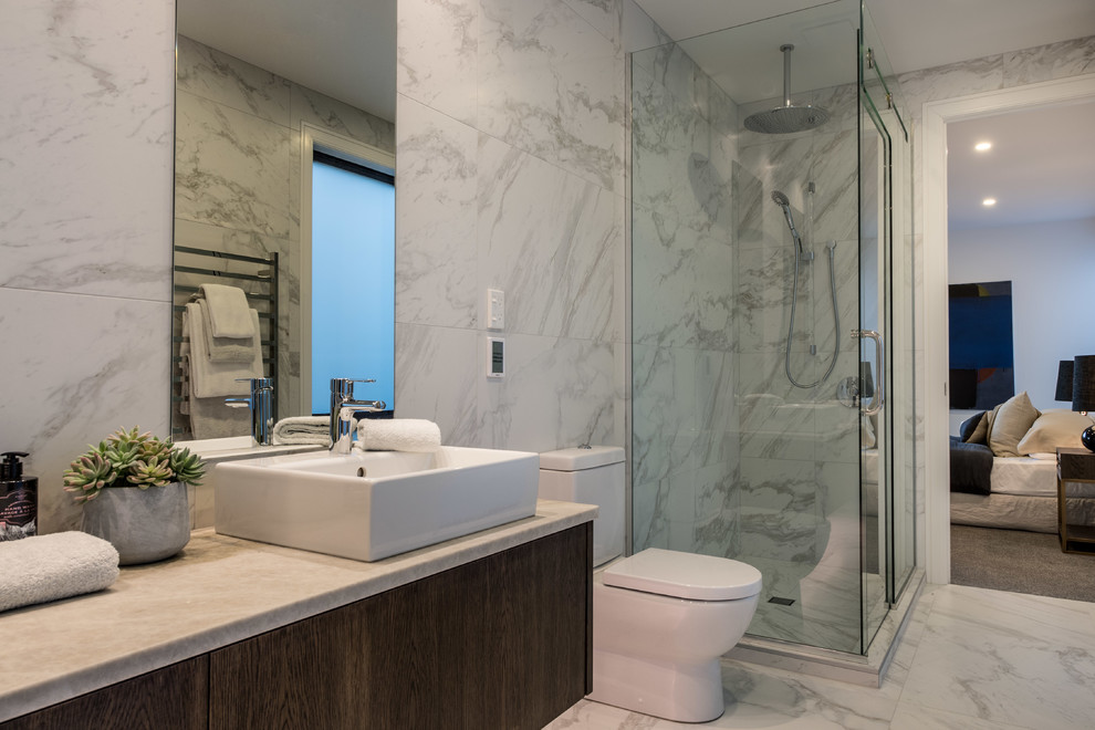 Design ideas for a medium sized contemporary ensuite bathroom in Auckland with medium wood cabinets, a freestanding bath, a corner shower, grey tiles, porcelain tiles, grey walls, porcelain flooring, a vessel sink, grey floors, a hinged door, flat-panel cabinets, a one-piece toilet and tiled worktops.