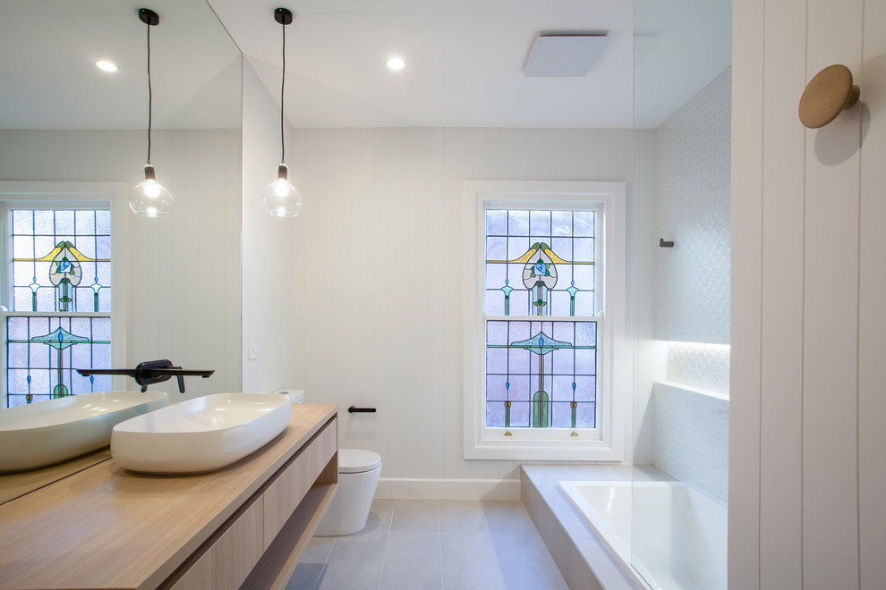 На фото: ванная комната в современном стиле с накладной ванной, белой плиткой, белыми стенами, серым полом, плоскими фасадами, светлыми деревянными фасадами, унитазом-моноблоком, душевой кабиной, настольной раковиной, столешницей из дерева и бежевой столешницей с