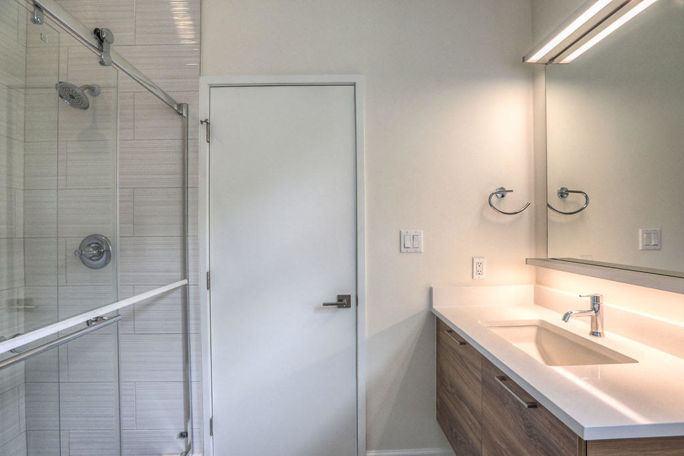 Bild på ett litet funkis badrum med dusch, med släta luckor, skåp i mellenmörkt trä, en dusch i en alkov, grå kakel, glaskakel, vita väggar, ett undermonterad handfat och marmorbänkskiva