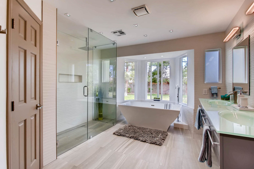 Идея дизайна: главная ванная комната в современном стиле с плоскими фасадами, серыми фасадами, отдельно стоящей ванной, угловым душем, белой плиткой, бежевыми стенами, монолитной раковиной, стеклянной столешницей, серым полом и душем с распашными дверями