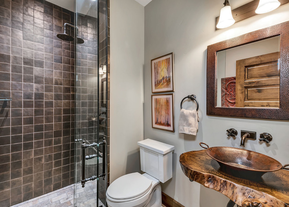 Rustik inredning av ett brun brunt badrum med dusch, med öppna hyllor, en dusch i en alkov, en toalettstol med separat cisternkåpa, brun kakel, grå väggar, ett fristående handfat, träbänkskiva och dusch med gångjärnsdörr