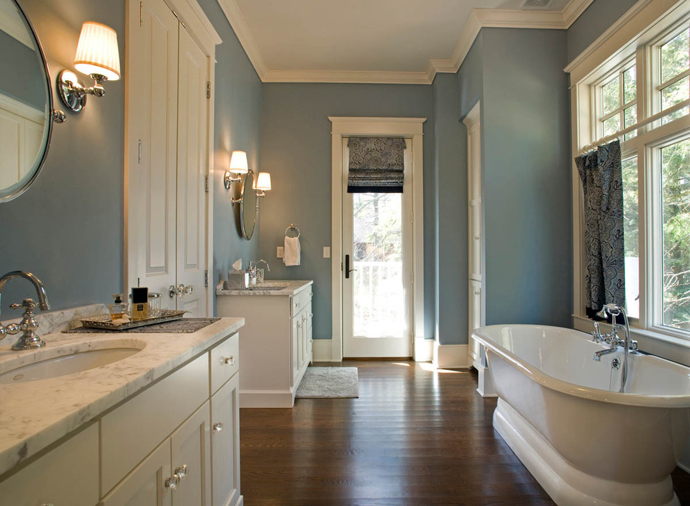 Réalisation d'une grande salle de bain principale tradition avec une baignoire indépendante, un placard avec porte à panneau encastré, des portes de placard blanches, un mur bleu, un sol en bois brun, un lavabo encastré, un plan de toilette en quartz et un sol marron.