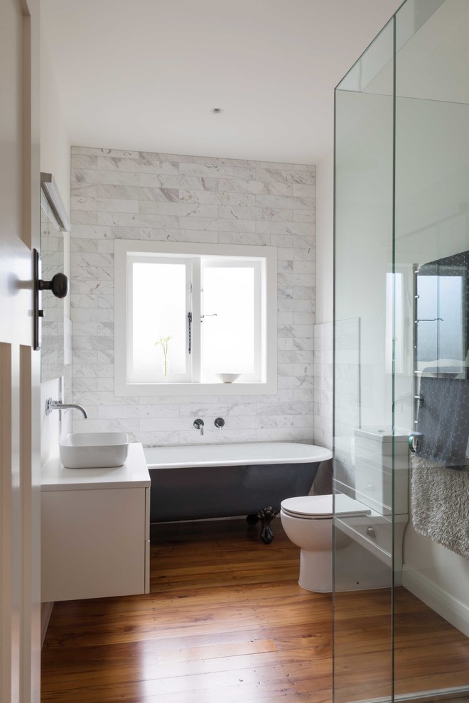 На фото: ванная комната в стиле неоклассика (современная классика) с белыми фасадами, ванной на ножках, белой плиткой, белыми стенами, паркетным полом среднего тона, настольной раковиной, коричневым полом и белой столешницей