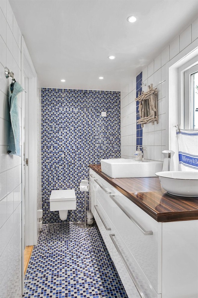 ストックホルムにある中くらいなビーチスタイルのおしゃれな浴室 (ベッセル式洗面器、フラットパネル扉のキャビネット、白いキャビネット、壁掛け式トイレ、青いタイル、モザイクタイル、モザイクタイル) の写真