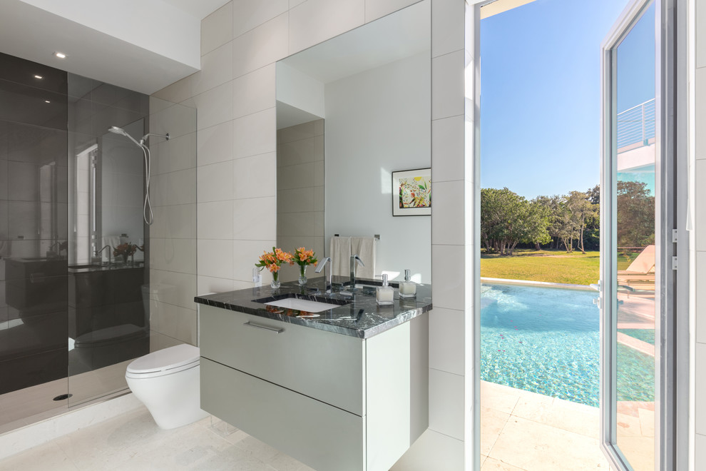 Photo of a medium sized contemporary bathroom in Tampa with flat-panel cabinets, grey cabinets, a freestanding bath, a wall mounted toilet, beige tiles, porcelain tiles, white walls, a submerged sink, marble worktops and an open shower.