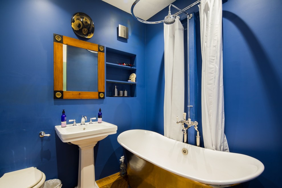 Photo of a medium sized modern family bathroom in London with a freestanding bath, a shower/bath combination, blue walls, a shower curtain, open cabinets, blue cabinets, a one-piece toilet, dark hardwood flooring, a pedestal sink and brown floors.