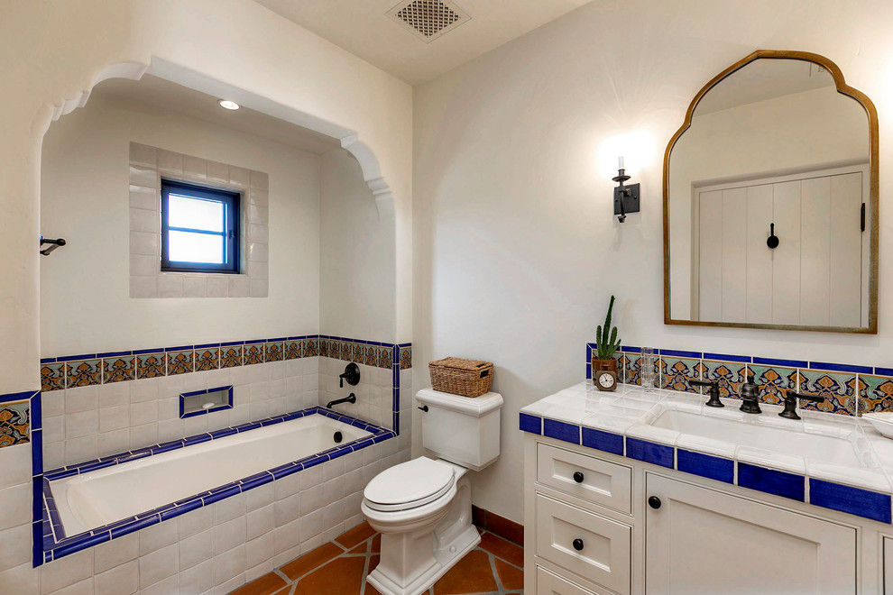 Foto di una stanza da bagno mediterranea di medie dimensioni con ante bianche, vasca ad alcova, WC a due pezzi, pareti bianche, pavimento in terracotta, lavabo sottopiano, top piastrellato e top bianco