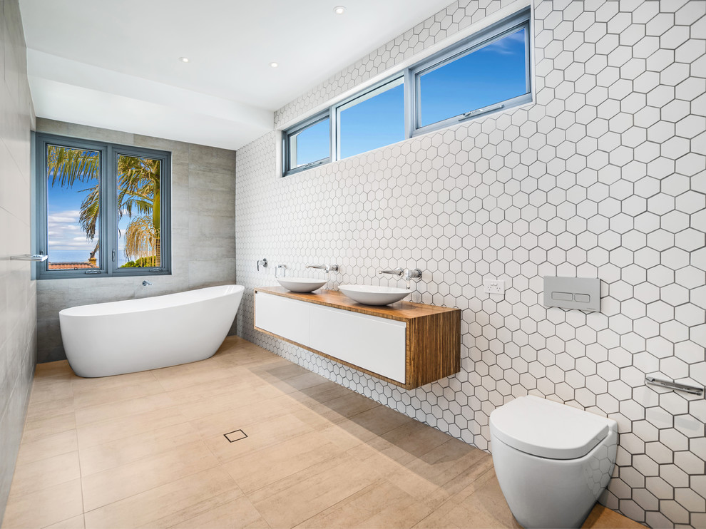 Foto på ett stort funkis brun badrum med dusch, med släta luckor, skåp i mellenmörkt trä, ett fristående badkar, en vägghängd toalettstol, vit kakel, ett fristående handfat, träbänkskiva och beiget golv