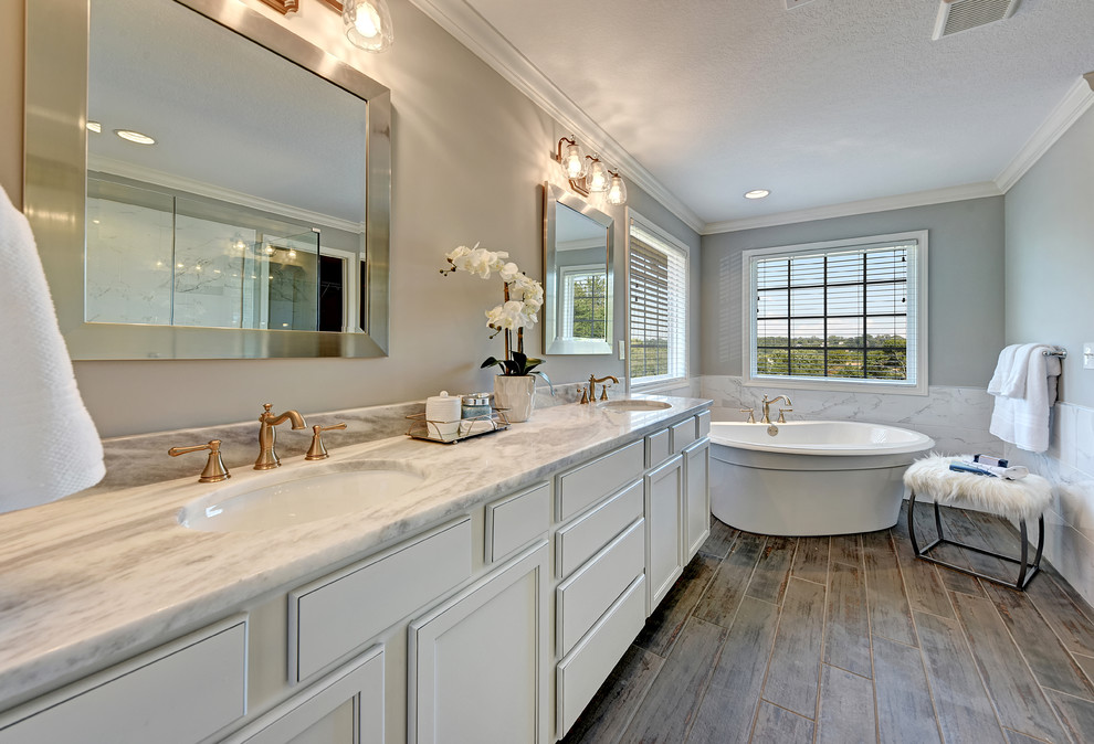 Foto de cuarto de baño clásico renovado con armarios con paneles empotrados, puertas de armario blancas, bañera exenta, losas de piedra, paredes grises, suelo de madera oscura, lavabo bajoencimera, suelo marrón y encimeras grises