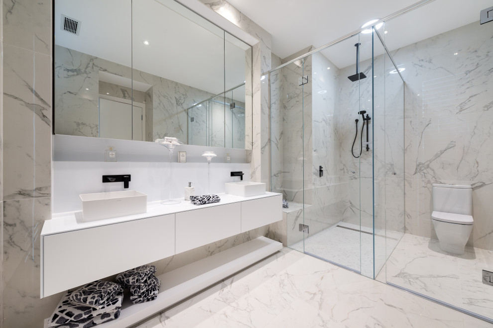 This is an example of a large contemporary ensuite bathroom in Melbourne with flat-panel cabinets, white cabinets, a corner shower, a one-piece toilet, grey tiles, a vessel sink, grey floors, a hinged door, black worktops, double sinks and a floating vanity unit.