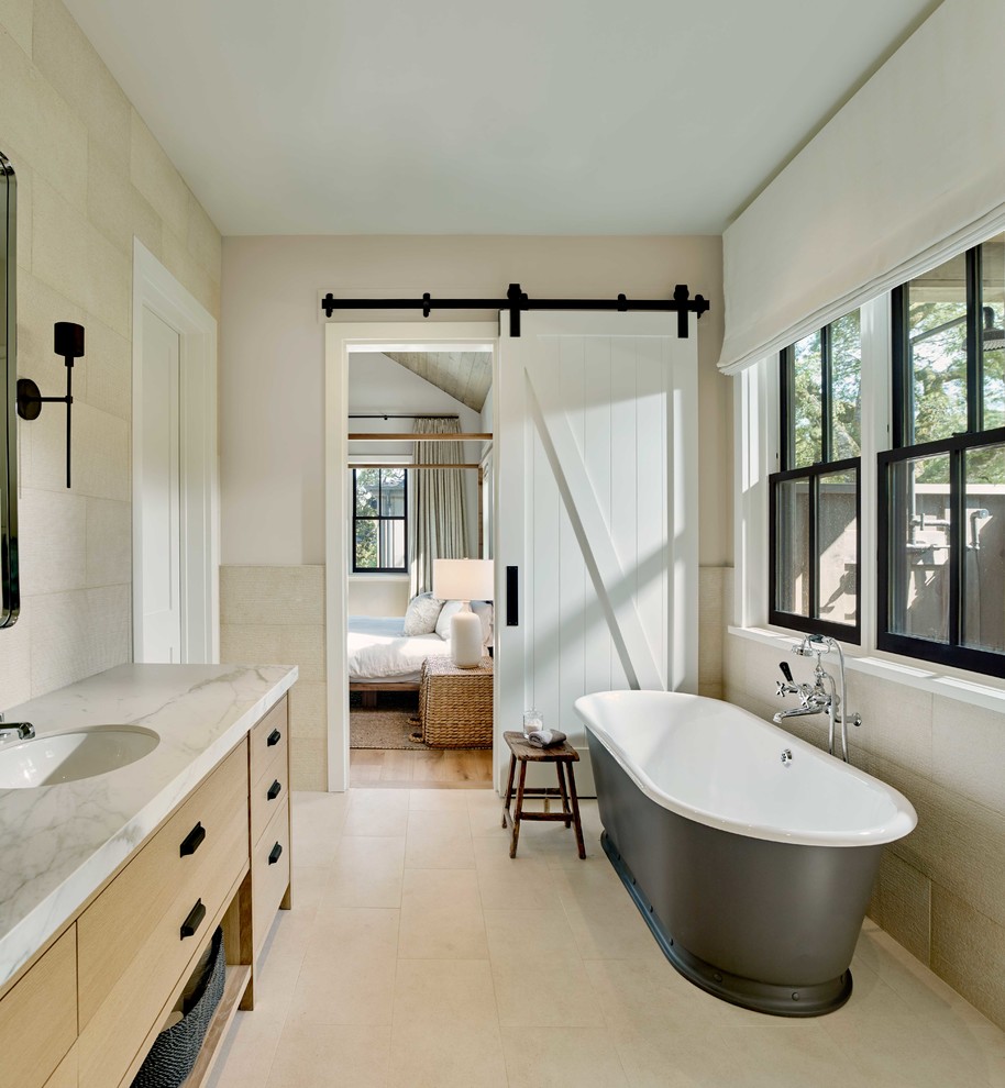 Réalisation d'une salle de bain principale champêtre en bois clair avec un placard à porte plane, une baignoire indépendante, un carrelage beige, un mur beige, un lavabo encastré, un sol beige et un plan de toilette blanc.