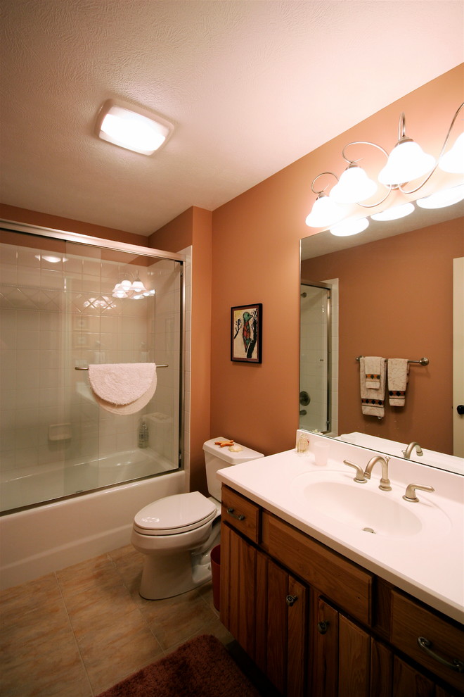 Foto på ett mellanstort amerikanskt badrum för barn, med ett integrerad handfat, luckor med upphöjd panel, skåp i mellenmörkt trä, bänkskiva i akrylsten, ett badkar i en alkov, en dusch/badkar-kombination, en toalettstol med separat cisternkåpa, beige kakel, keramikplattor, orange väggar och travertin golv