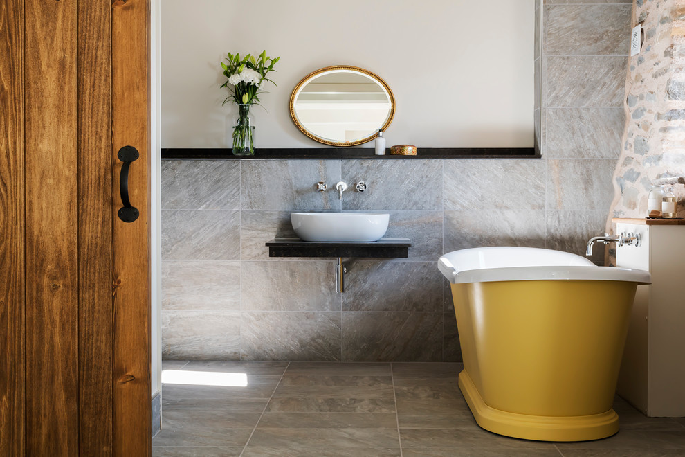 Cette image montre une grande salle de bain principale et grise et jaune rustique avec une baignoire indépendante, un carrelage gris, un carrelage de pierre, un mur gris, une vasque, un plan de toilette en marbre et un sol gris.