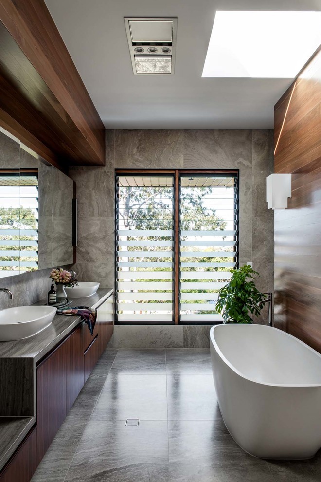 Пример оригинального дизайна: ванная комната в стиле модернизм с плоскими фасадами, темными деревянными фасадами, отдельно стоящей ванной, серой плиткой, настольной раковиной, серым полом и серой столешницей